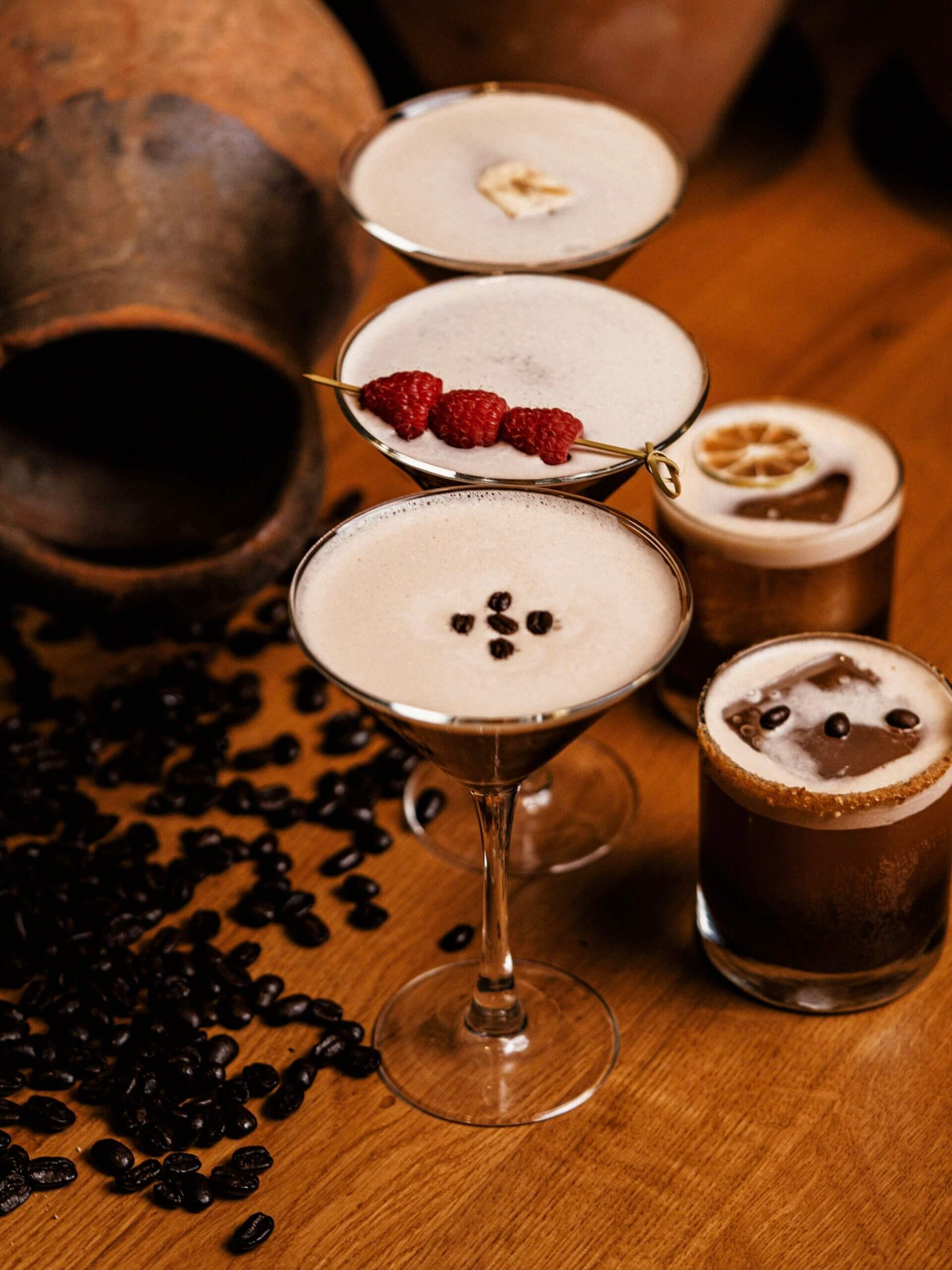 Fem stycken olika kaffedrinkar med tre höga cocktailglas och två whiskeyglas och kaffebönor runt bordet 