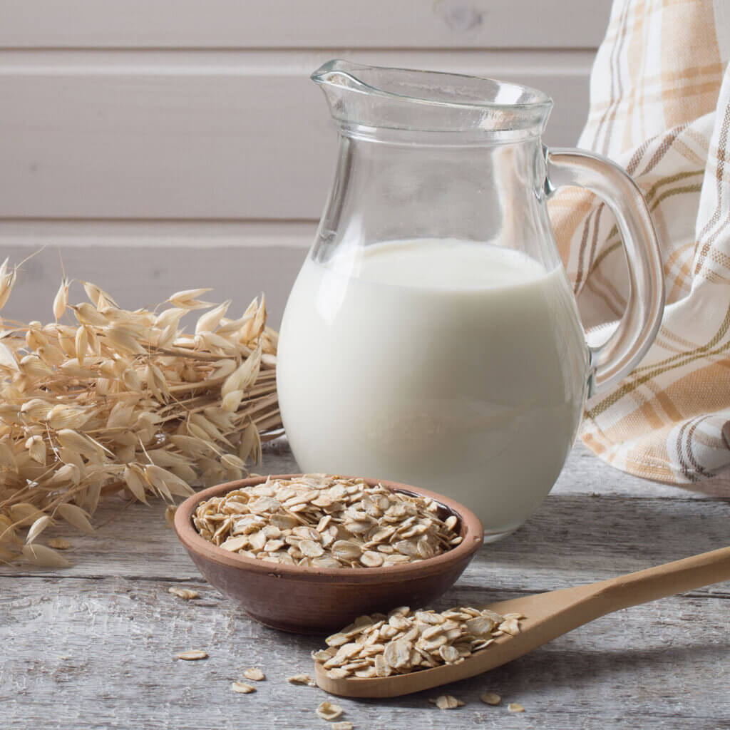 En kanna med havremjölk. Framför står en skål och en sked med havregryn. Bakom ligger ett havre-ax.